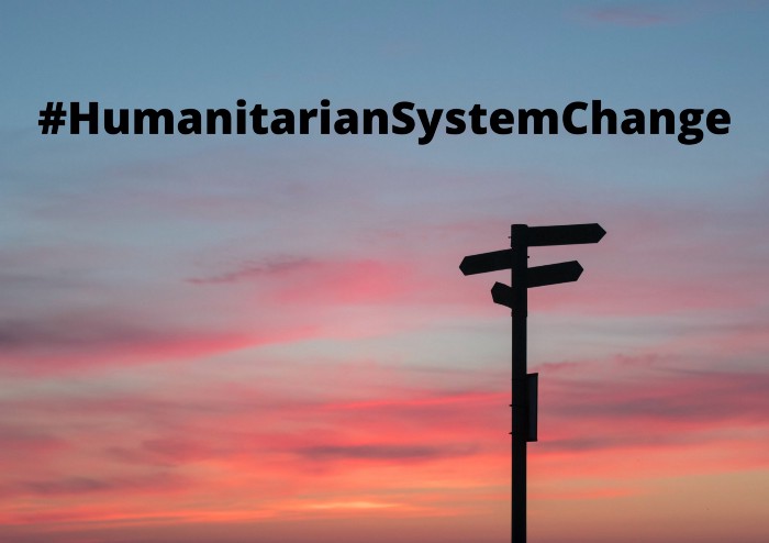 A signpost is silhouetted against a sunset. #HumanitarianSystemChange is written across the sky.